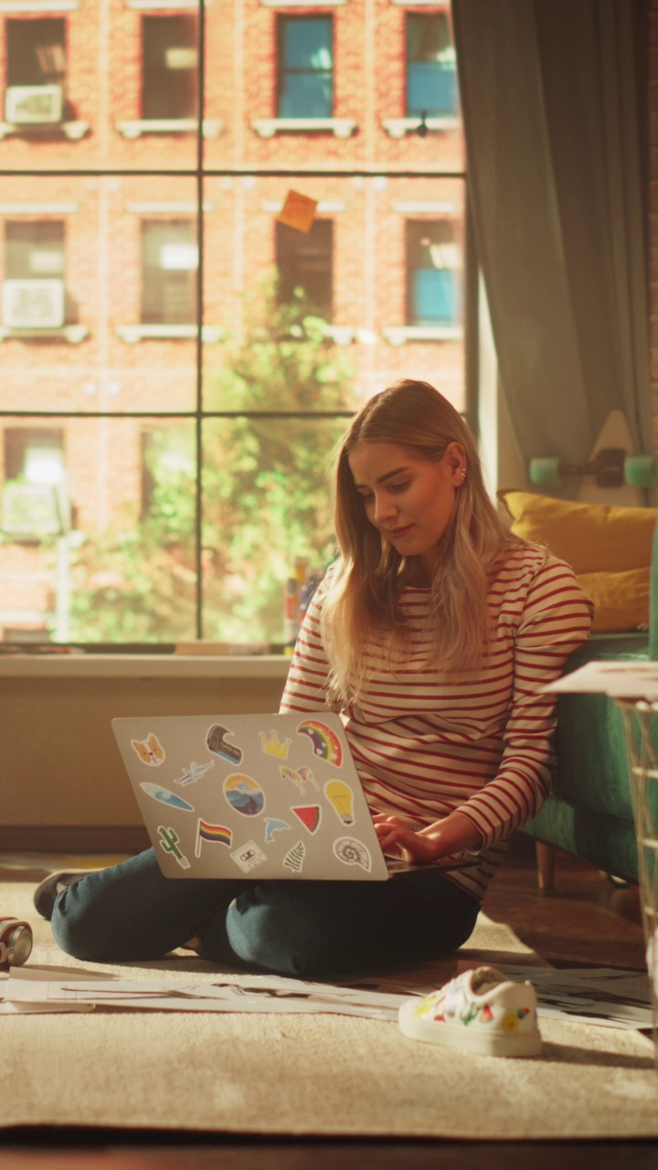 垂直视角的金发女人使用笔记本电脑，而坐在客厅地板上舒适时尚的公寓。迷人的女孩在思考和编码视频下载