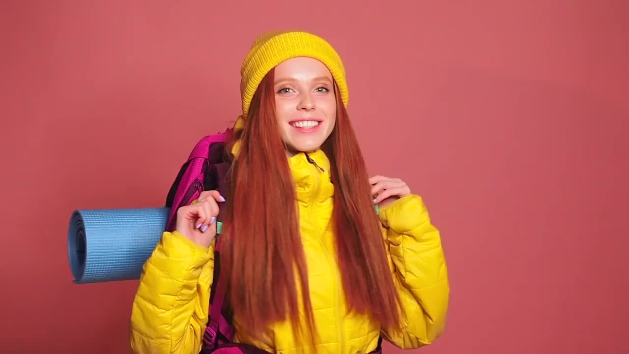 红发姜黄色的女人在粉红色的工作室背景穿着黄色的风衣夹克和帽子，在她的肩膀上的背包。她准备走了视频下载
