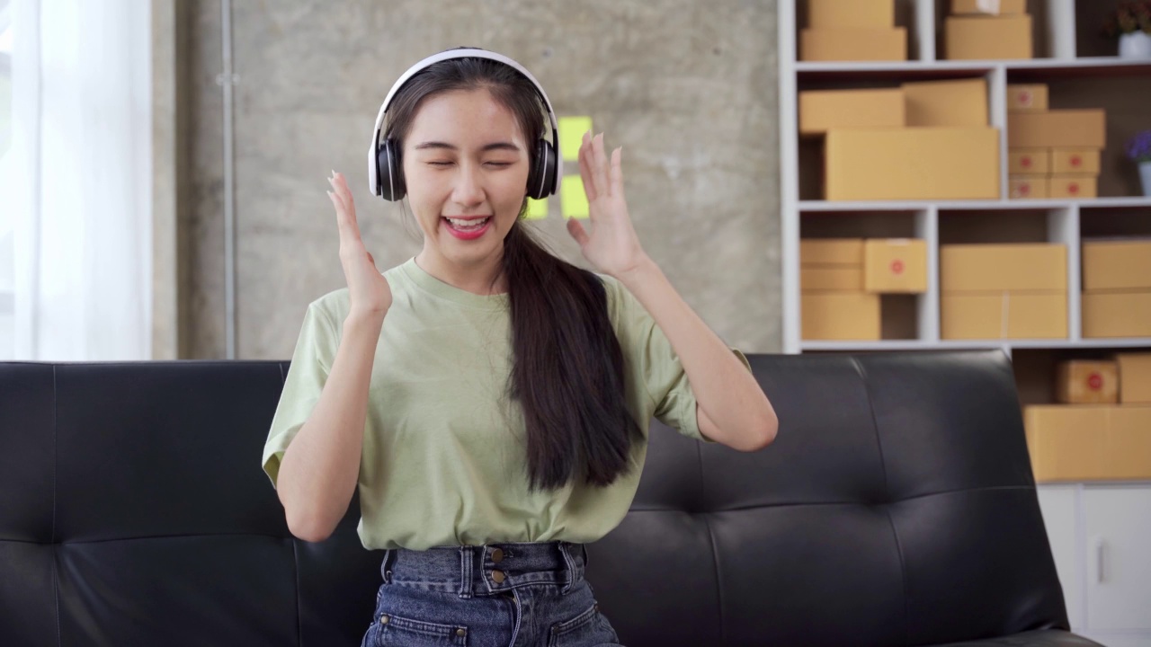 有魅力的亚洲女性在家里享受音乐，戴着耳机，拿着智能手机，在沙发上唱歌跳舞。现代科技和年轻人的观念。视频素材