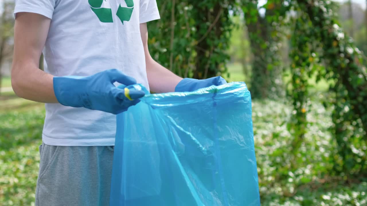 在一个被污染的公园里，两名男子用塑料袋收集塑料垃圾。橡胶手套，t恤上的回收标志视频素材
