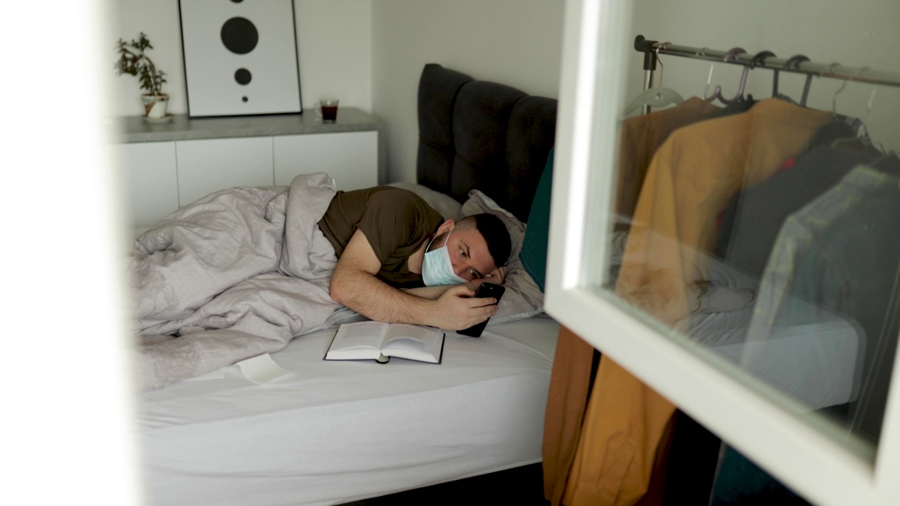 冠状病毒大流行期间，一名年轻人在床上使用智能手机并戴着外科口罩视频素材