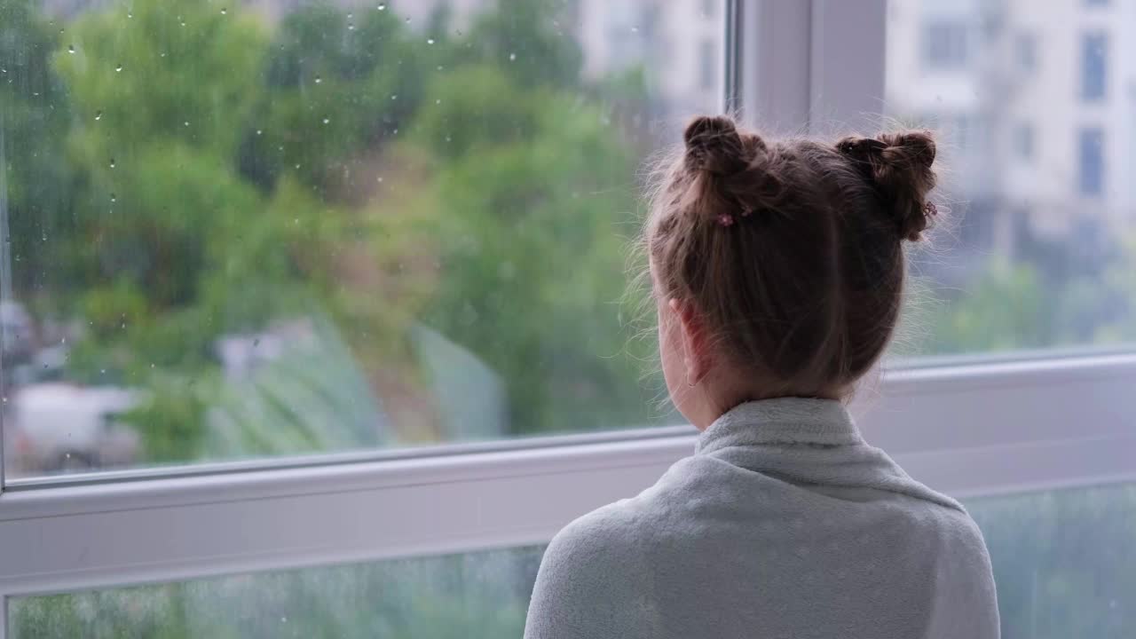 女孩看着窗外的雨。因为天气不好，靠窗的孩子很伤心。视频素材