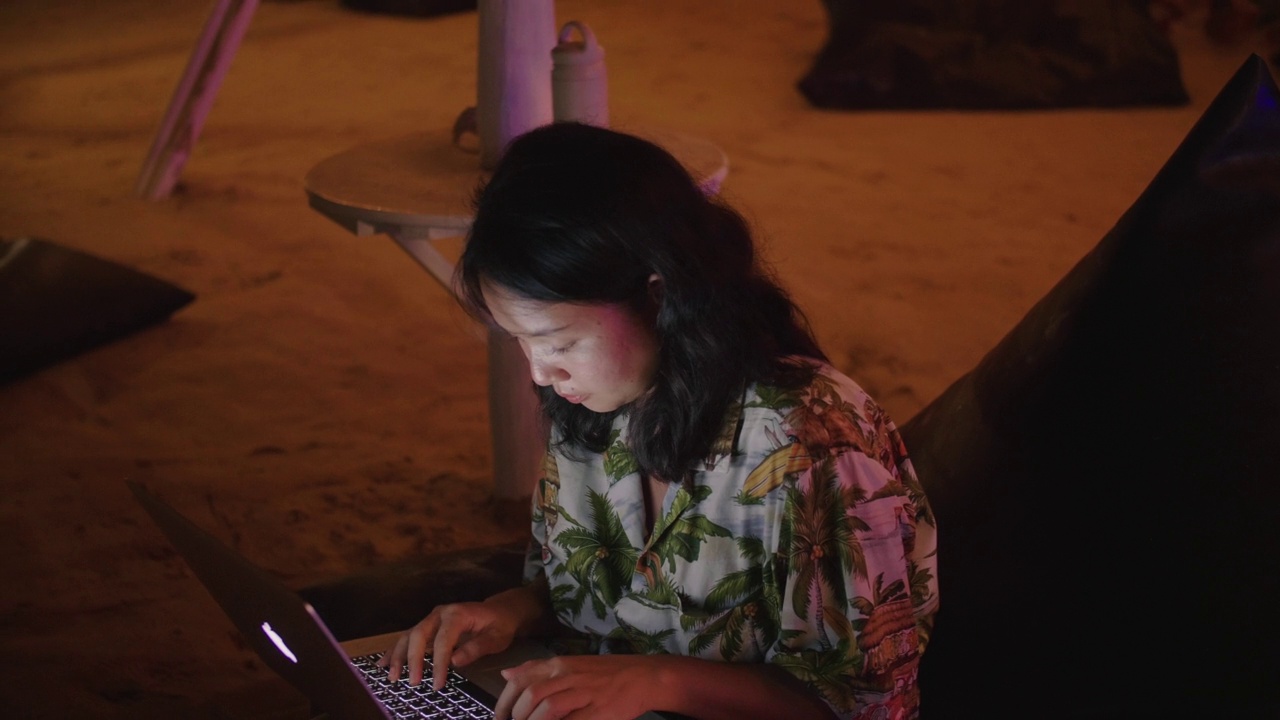 年轻的亚洲女游客坐在海滩附近，晚上在海边使用笔记本电脑。视频素材