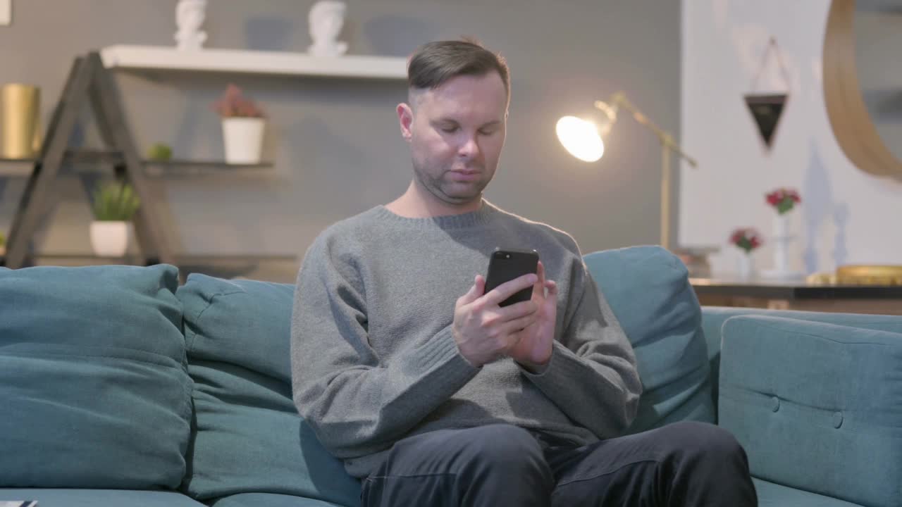 休闲男人坐在沙发上玩智能手机视频下载