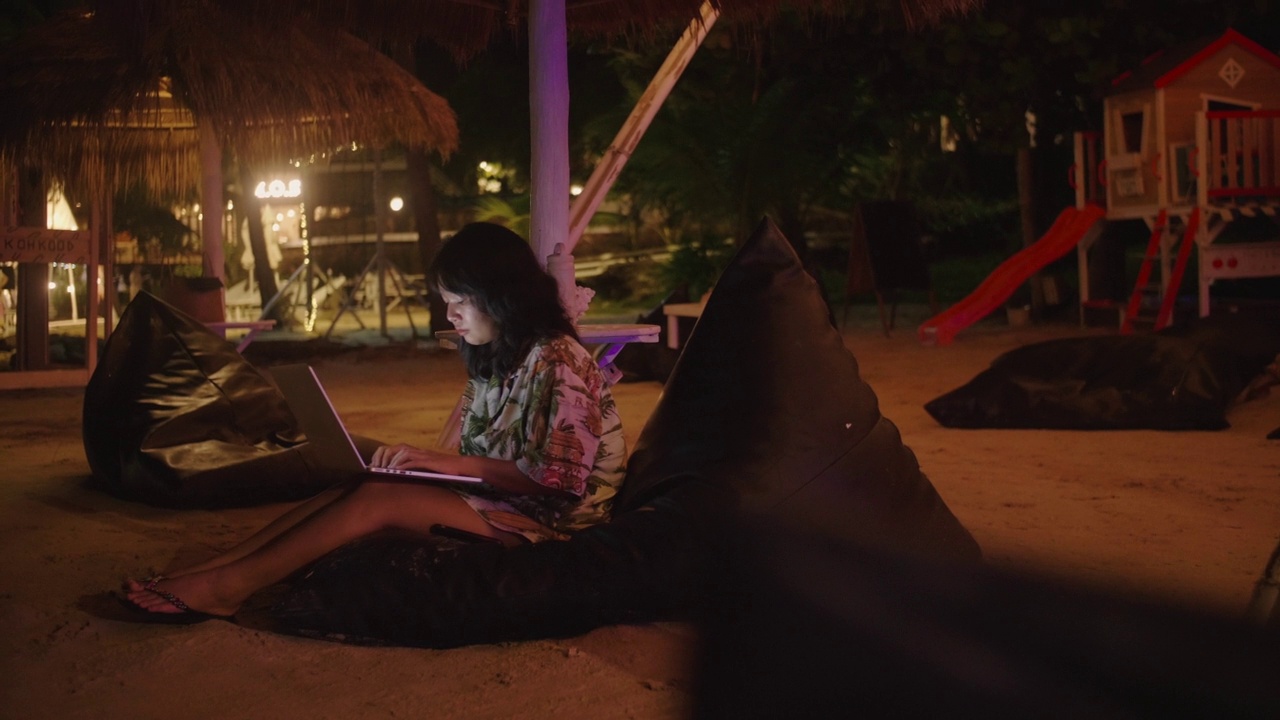 年轻的亚洲女性在海边的夜晚，在键盘上打字，使用笔记本电脑的触控板享受假期。视频素材