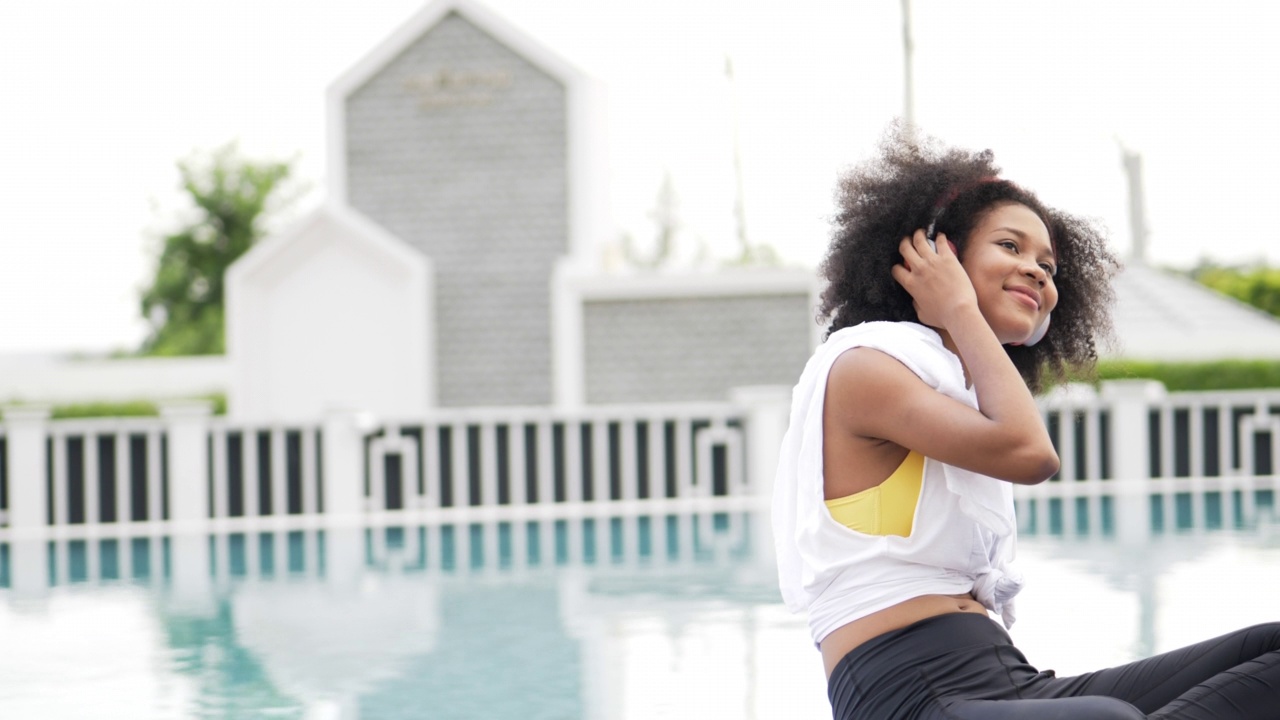 穿着运动服的非洲妇女戴着耳机在游泳池的背景。放松喜欢听轻松的音乐。股票视频视频素材