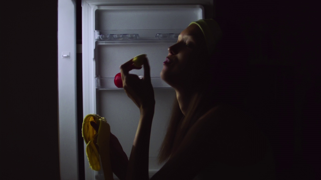 一名饥饿的年轻女子在晚上打开冰箱，坐在地板上吃东西的特写。视频下载