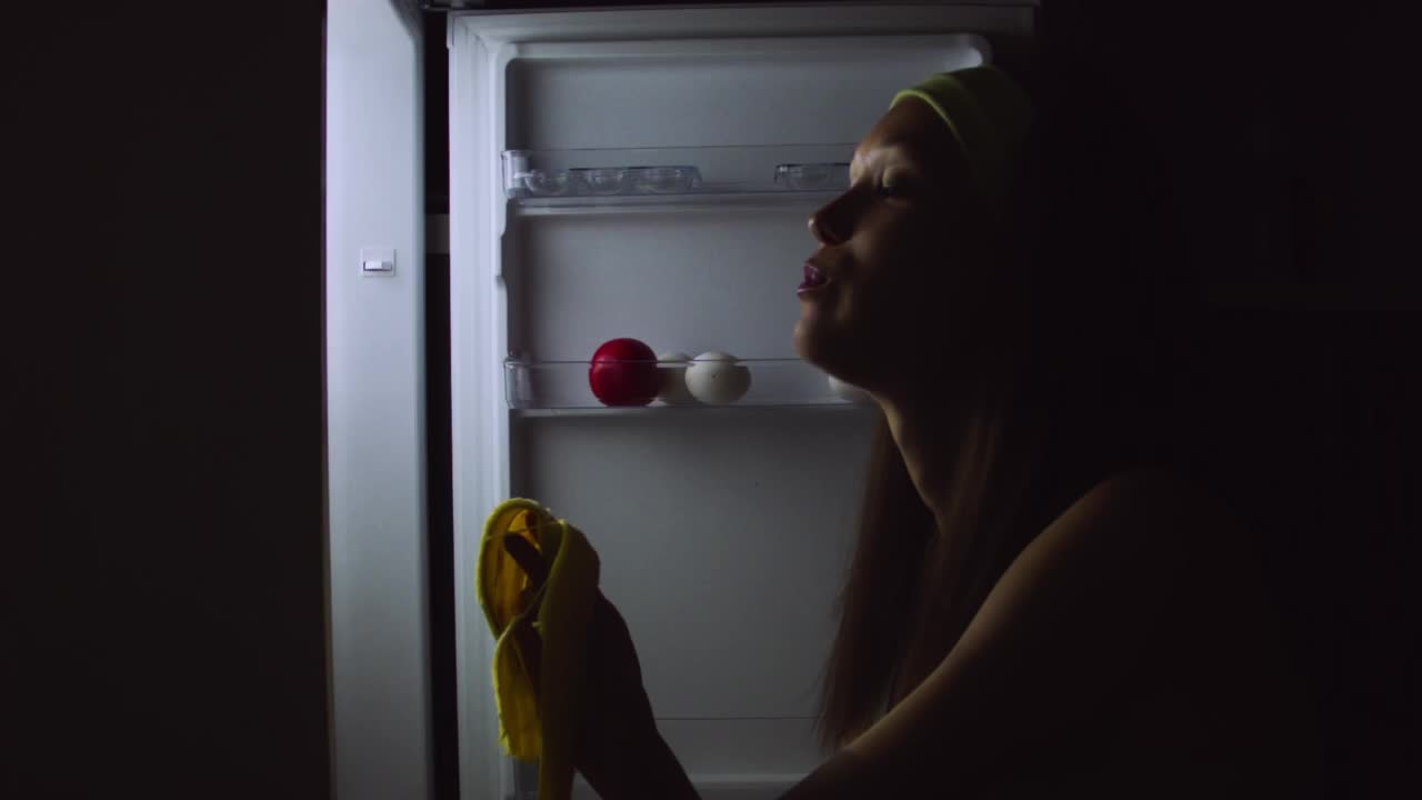 一名饥饿的年轻女子在晚上打开冰箱，坐在地板上吃东西的特写。视频下载
