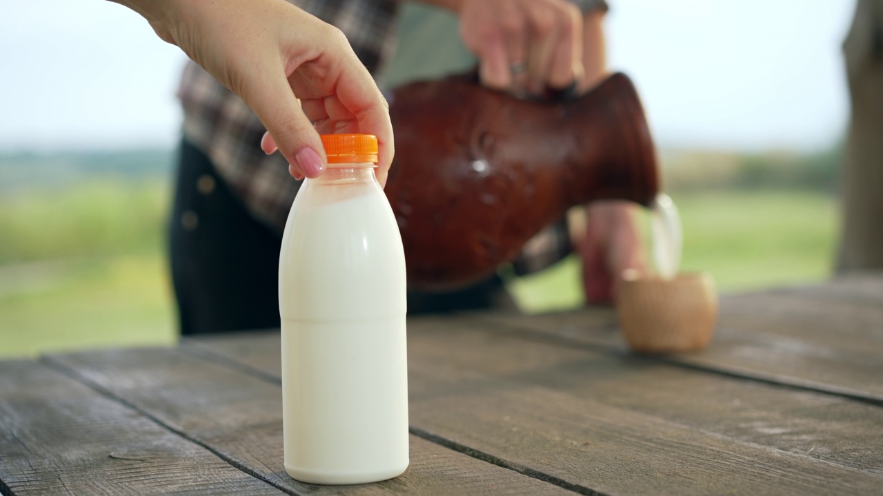 模糊的难以辨认的男人倒牛奶从陶罐在杯子与女性的手把奶瓶放在桌子在前面。农田户外健康乳制品广告。视频下载