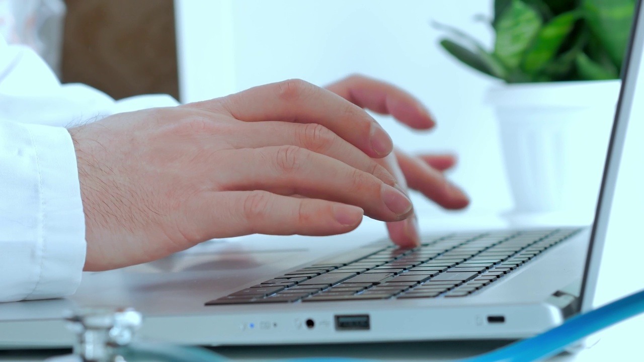 医学的概念，医生的工作，一个穿着白袍的男人的手在笔记本电脑上打印文本，在白色的桌子上一个风扇内窥镜和一株休眠的绿色植物视频素材