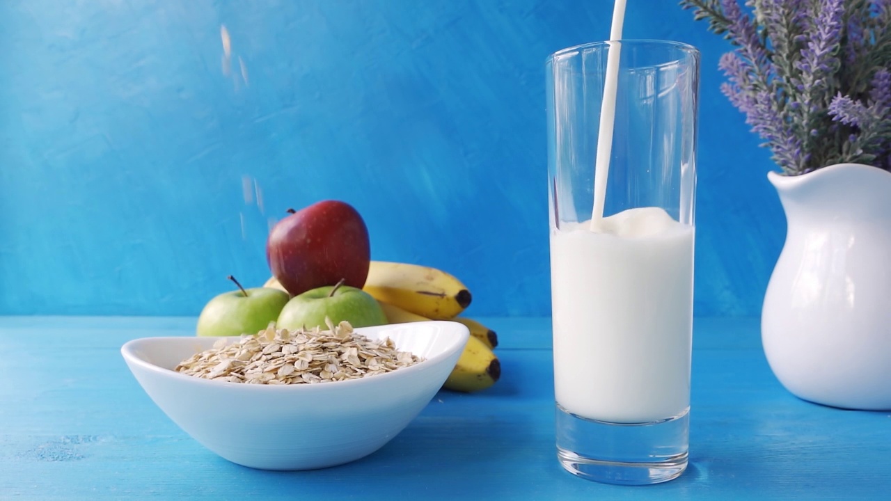 牛奶倒进玻璃杯，燕麦片倒进碗里，在蓝色的木桌上放着新鲜的苹果和香蕉。4k原始视频60帧/秒。视频素材
