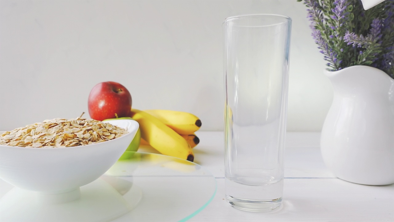 牛奶倒入一杯，燕麦片饮食谷类在一个碗旋转玻璃托盘上的白色木桌新鲜的绿色和红色的苹果和香蕉。4k原始视频60帧/秒。视频素材