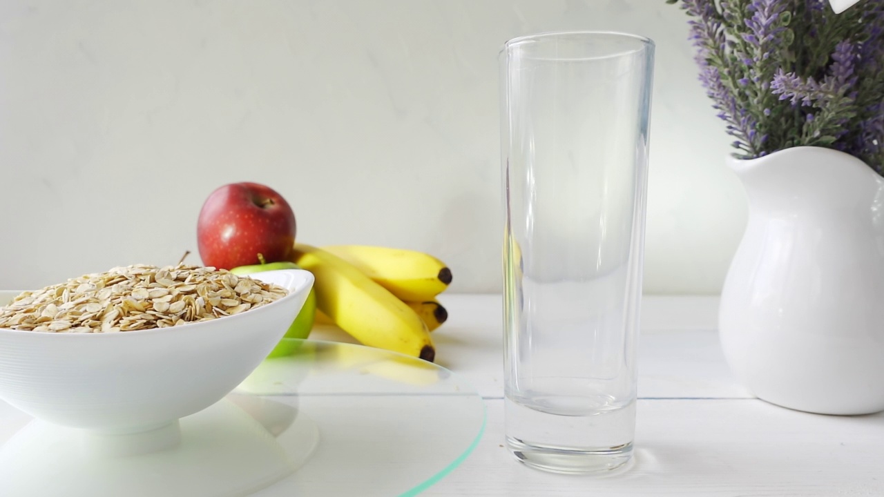 牛奶倒入一杯，燕麦片饮食谷类在一个碗旋转玻璃托盘上的白色木桌新鲜的绿色和红色的苹果和香蕉。4k原始视频60帧/秒。视频素材