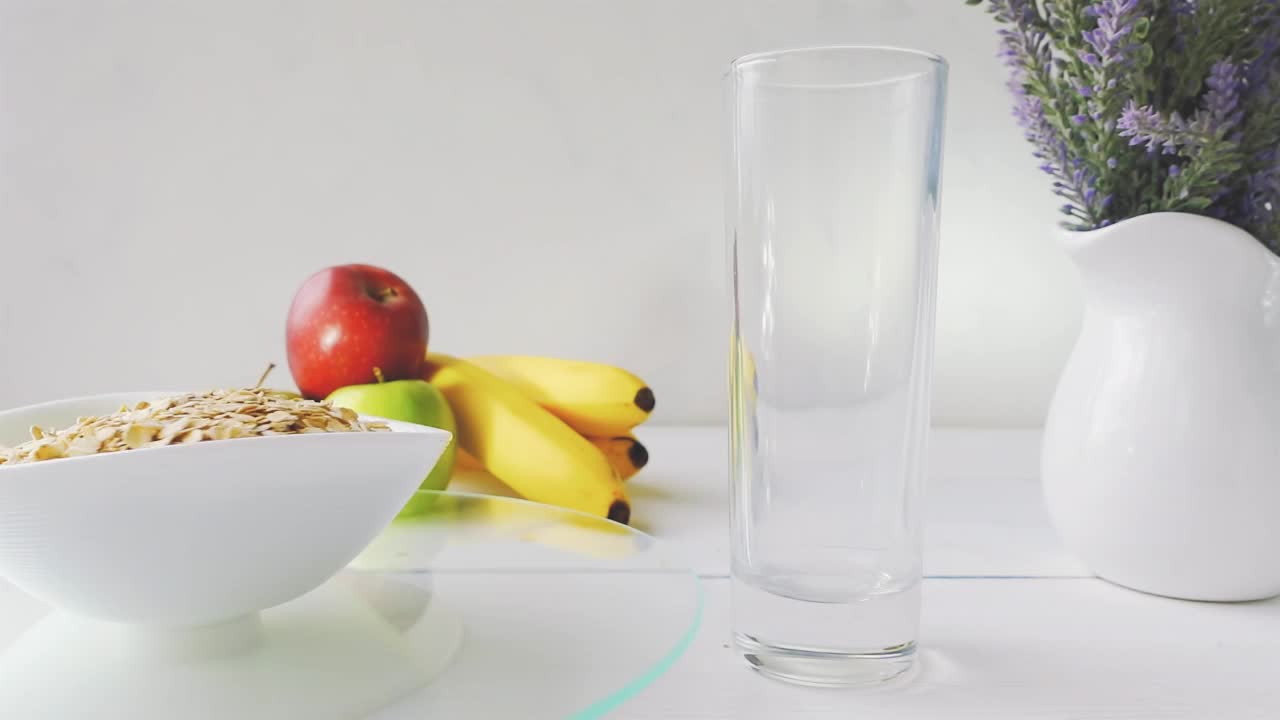 牛奶倒入一杯，燕麦片饮食谷类在一个碗旋转玻璃托盘上的白色木桌新鲜的绿色和红色的苹果和香蕉。带有速度斜坡效果的4k原始视频。视频素材