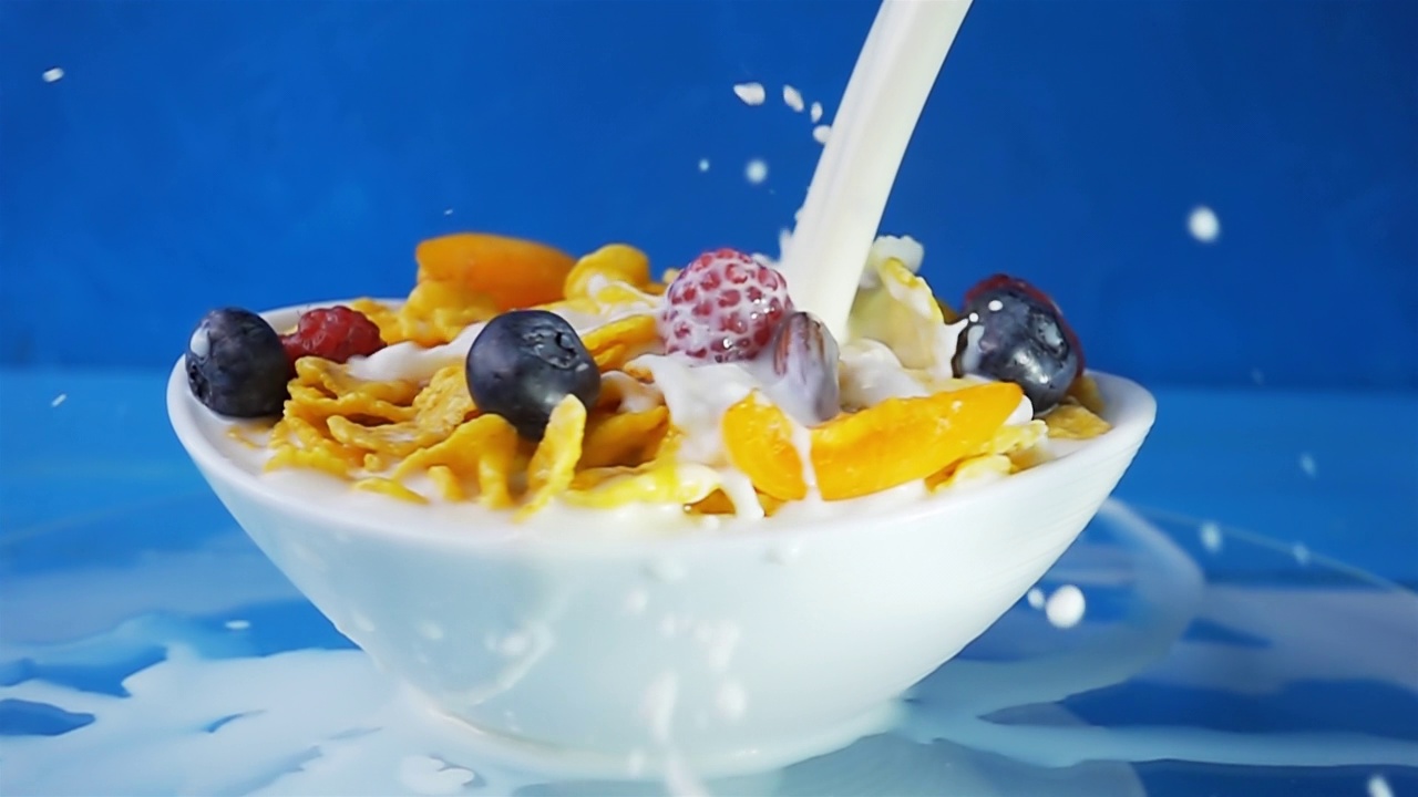 牛奶倒进黄色的玉米片里，配上各种新鲜的蓝莓、覆盆子、葡萄和杏仁，放在一个浅蓝色背景的白色旋转盘里。4k原始慢动作视频。视频素材