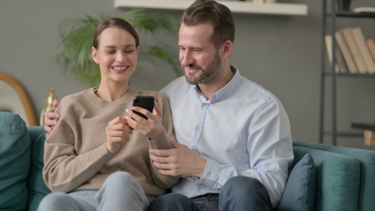 情侣在智能手机上庆祝成功的肖像，坐在沙发上视频素材