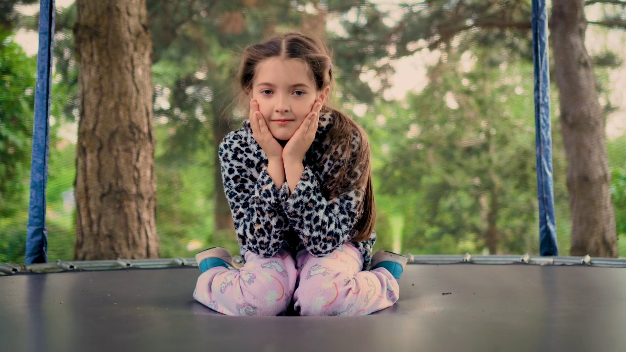 肖像可爱微笑的女孩坐在蹦床在家里的花园视频素材