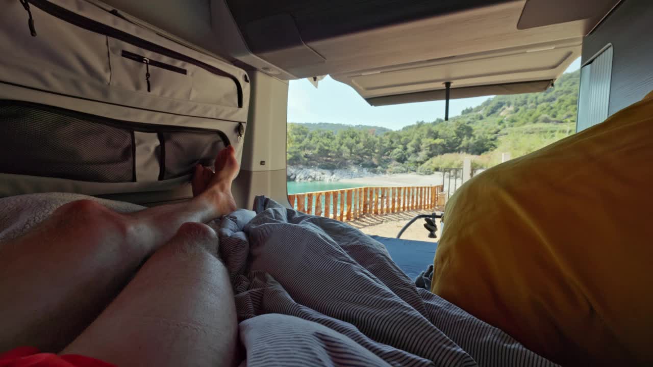 男人躺在他的面包车的床上，面包车的后门开着，美丽的海滩在后面视频下载