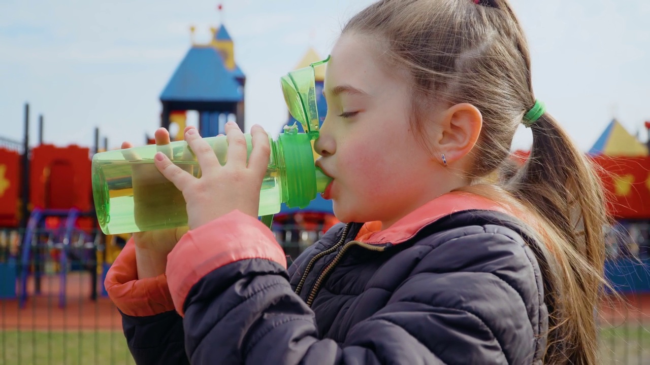 在娱乐公园背景的儿童游乐场上，小女孩正在喝瓶装水视频素材