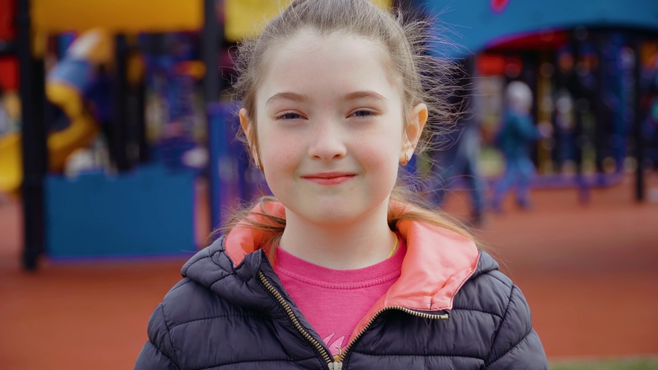 在娱乐公园背景的儿童游乐场上，可爱的小女孩看着镜头微笑的肖像视频素材