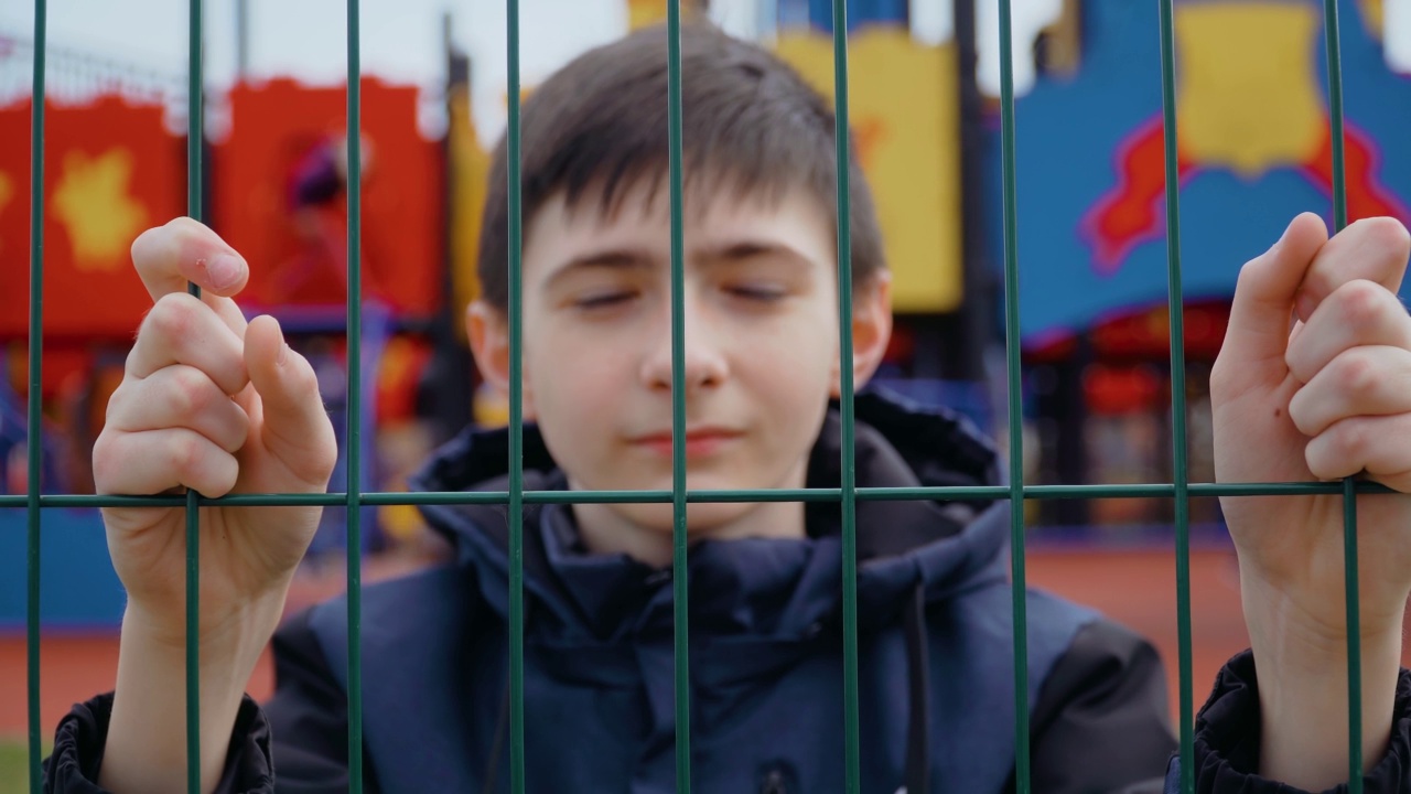 伤心难过的男孩在金属格栅后面看着相机的肖像在娱乐公园与儿童游乐场的背景视频素材
