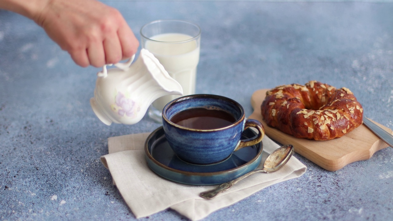 一名妇女用牛奶壶将牛奶倒入一杯咖啡中。健康早餐咖啡加牛奶和面包。4 k的决议视频下载