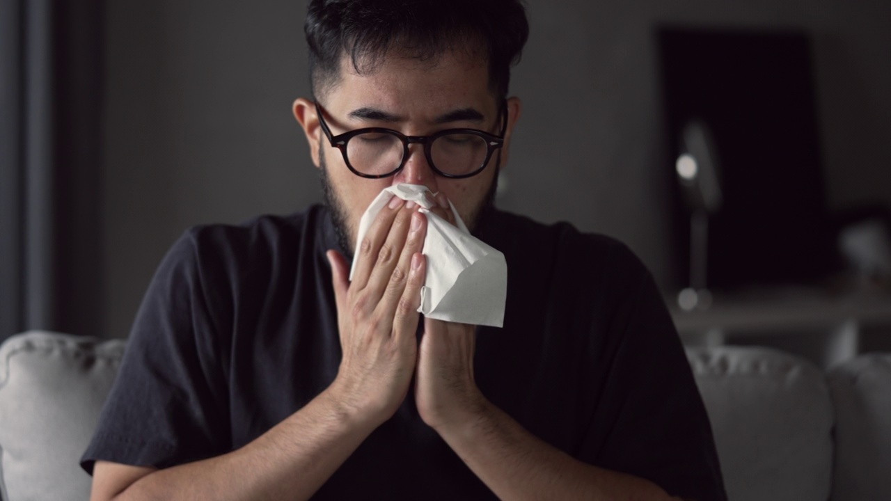 亚洲男子生病了，在家里的沙发上用纸巾打喷嚏。男性患感冒擤鼻涕。寒热概念。视频素材