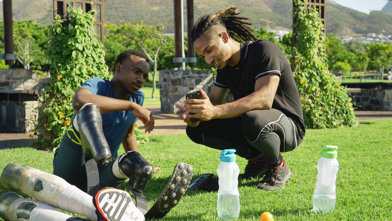 男性截肢运动员和教练在公园草地上使用智能手机视频素材