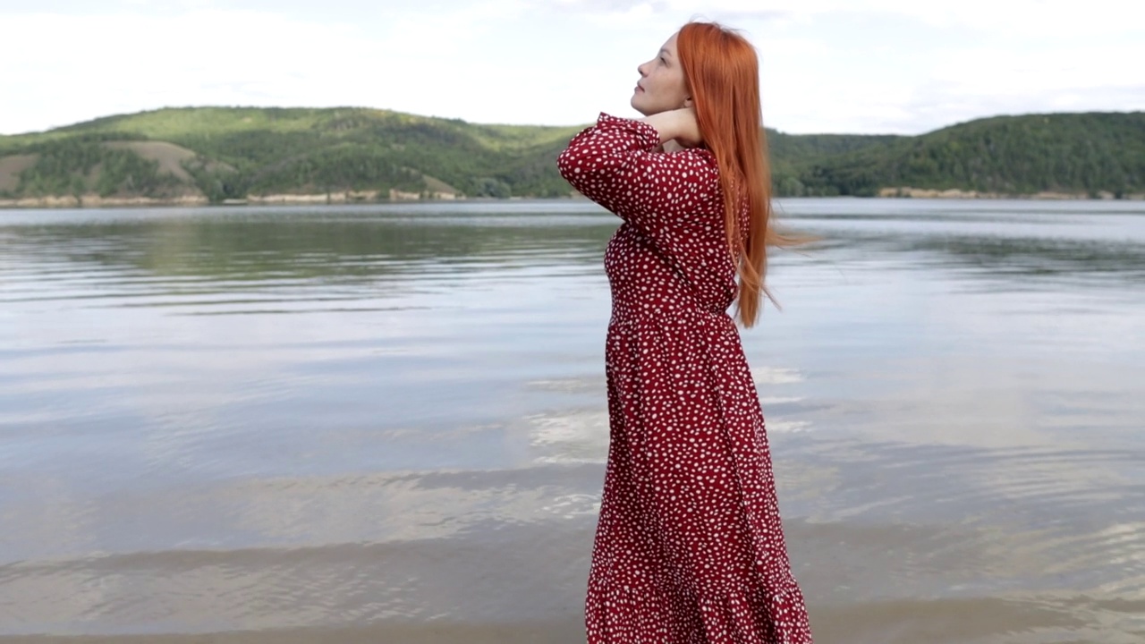 年轻的红头发，美丽的女人穿着红色的裙子赤脚走在山湖的沙滩上视频素材