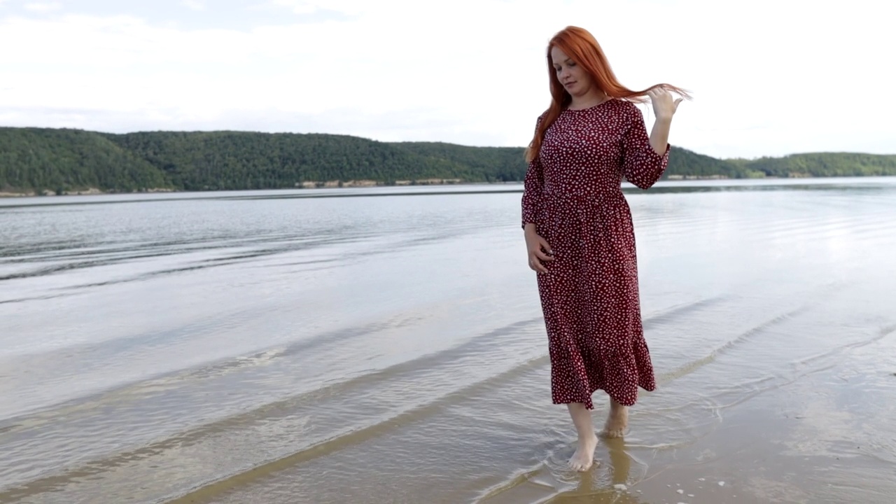 年轻的红头发，美丽的女人穿着红色的裙子赤脚走在山湖的沙滩上视频素材