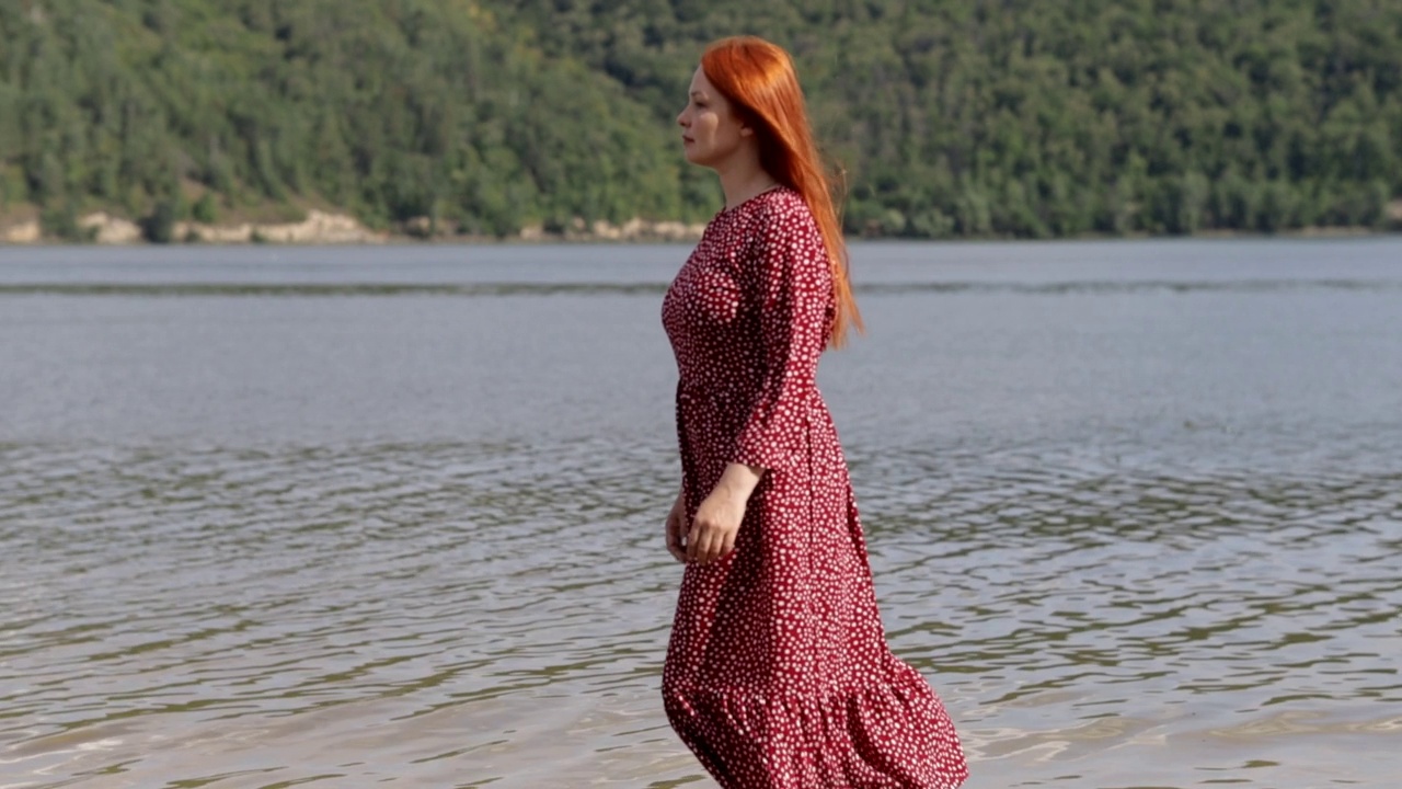 年轻的红头发，美丽的女人穿着红色的裙子走在山湖的沙滩上视频素材