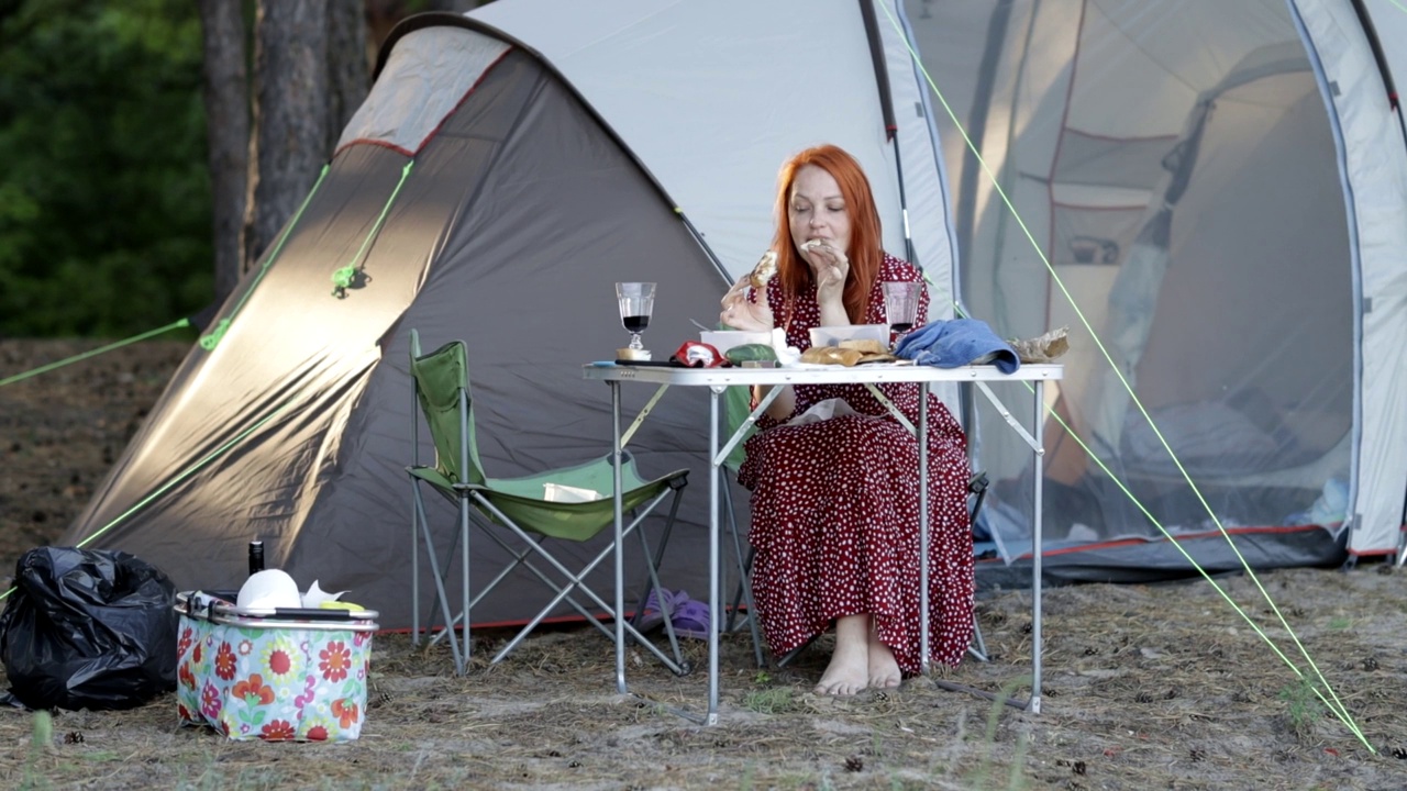 年轻的红发女人在松林吃牛排，帐篷旁露营。视频素材