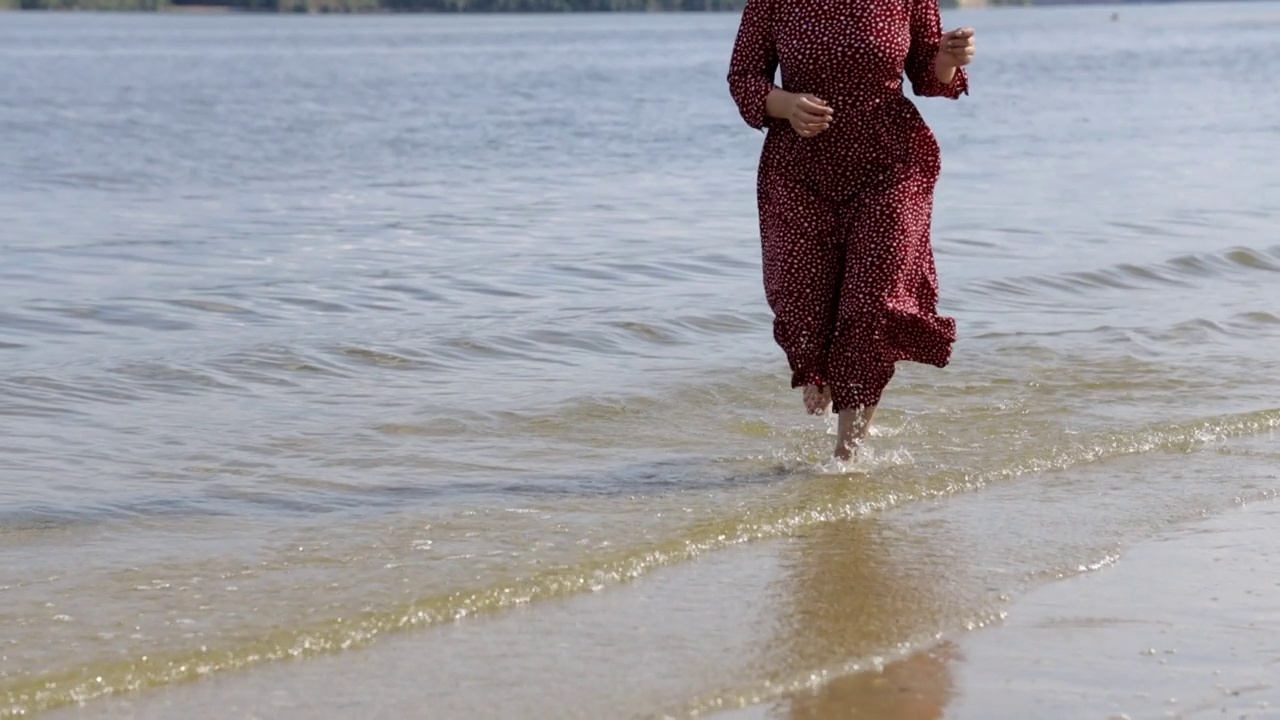 快乐的年轻红头发，美丽的女人穿着红裙子沿着山中的沙滩跑视频素材