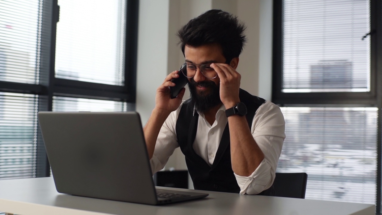 成功的胡子自由职业者男性，戴着眼镜在笔记本电脑前工作，坐在桌子上，通过手机与同事讨论项目细节。视频素材