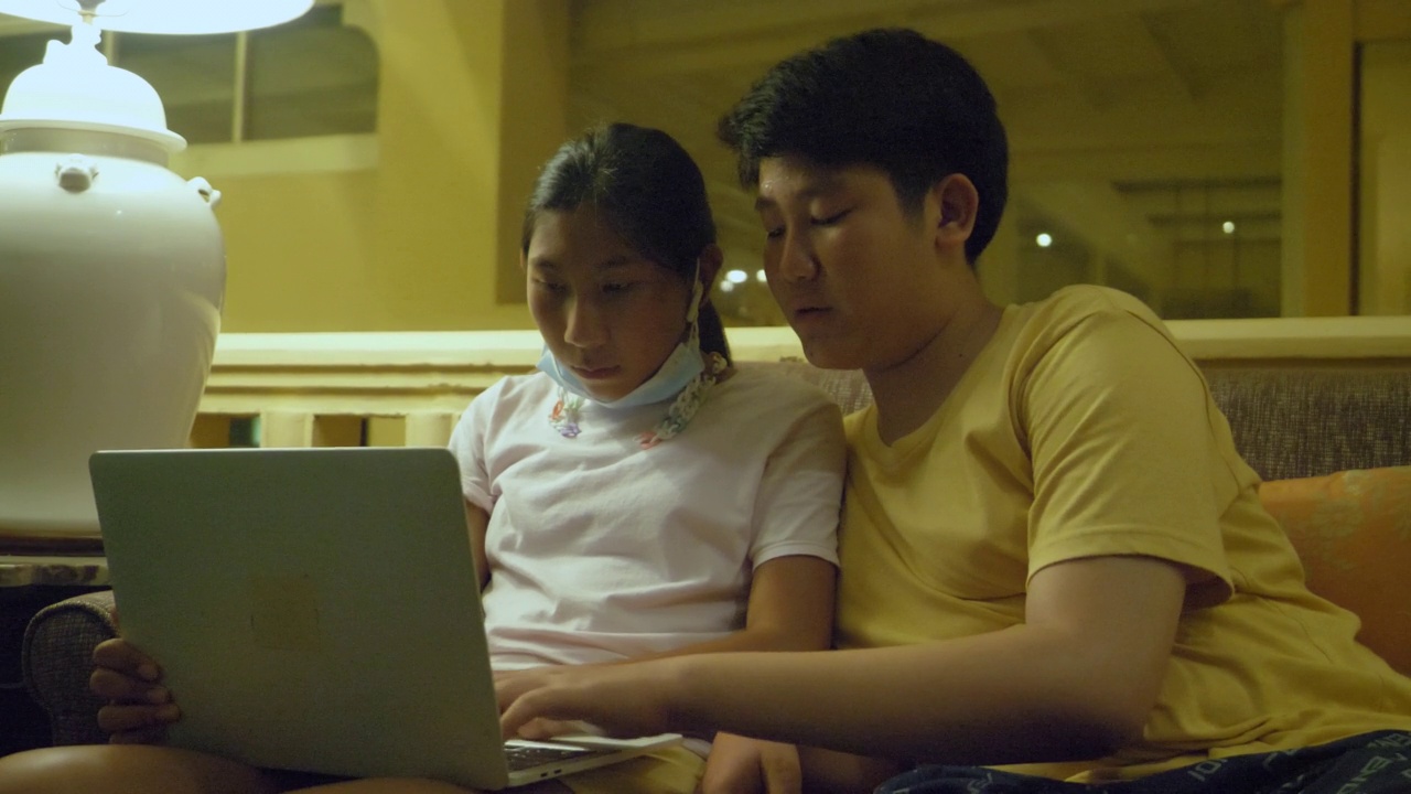 亚洲男孩在度假期间在酒店大堂的沙发上教他的妹妹使用笔记本电脑。视频素材