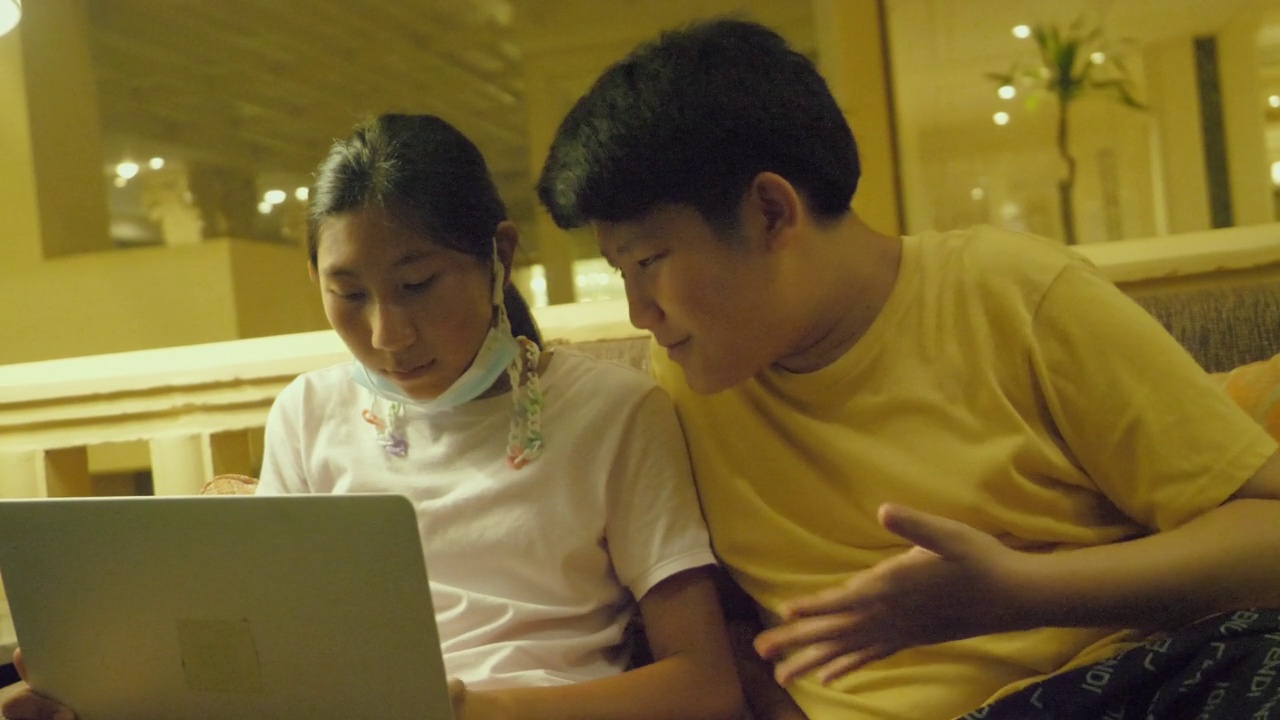 亚洲男孩在度假期间在酒店大堂的沙发上教他的妹妹使用笔记本电脑。视频素材