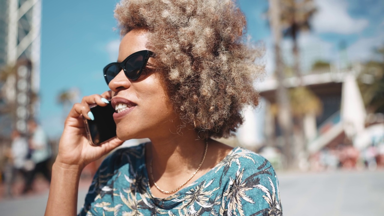 微笑的非洲灰色头发的女人戴着墨镜在长凳上通过手机交谈视频素材