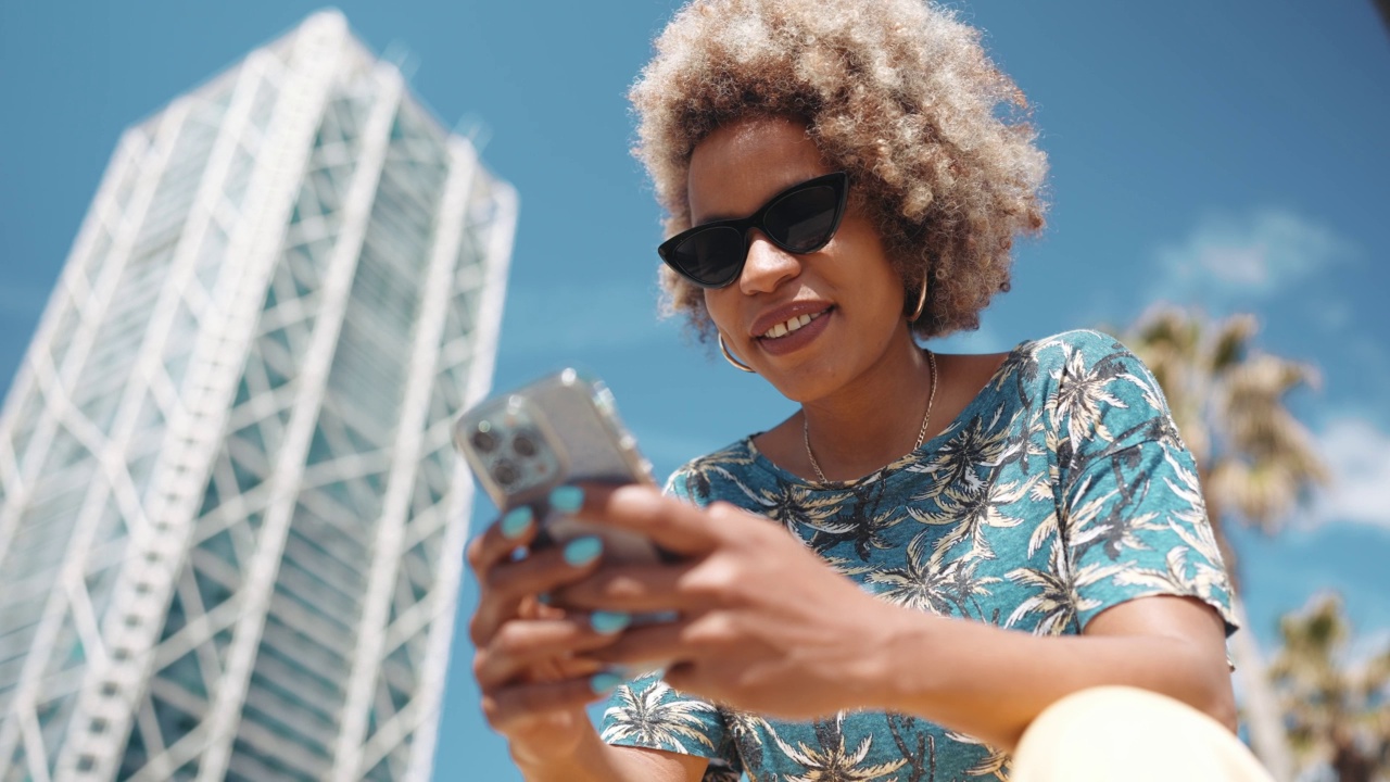 高兴的非洲灰色头发的女人戴着太阳镜在长凳上用手机发短信视频素材