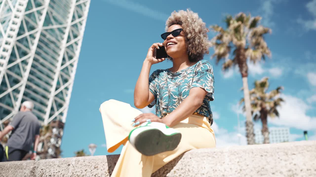 笑着的非洲灰色头发的女人戴着墨镜在长凳上用手机说话视频素材