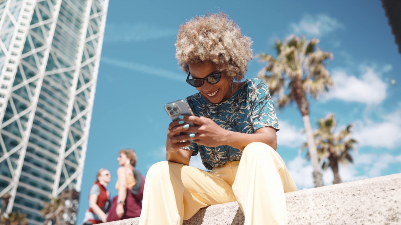 微笑的非洲灰色头发的女人戴着墨镜在长凳上用手机发短信视频素材