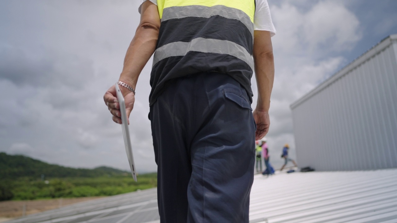 真实的亚洲男性工程师或技术人员步行到工厂屋顶上工作，利用太阳能发电，手持数字平板检查太阳能电池板的运行和检查维护系统。视频下载