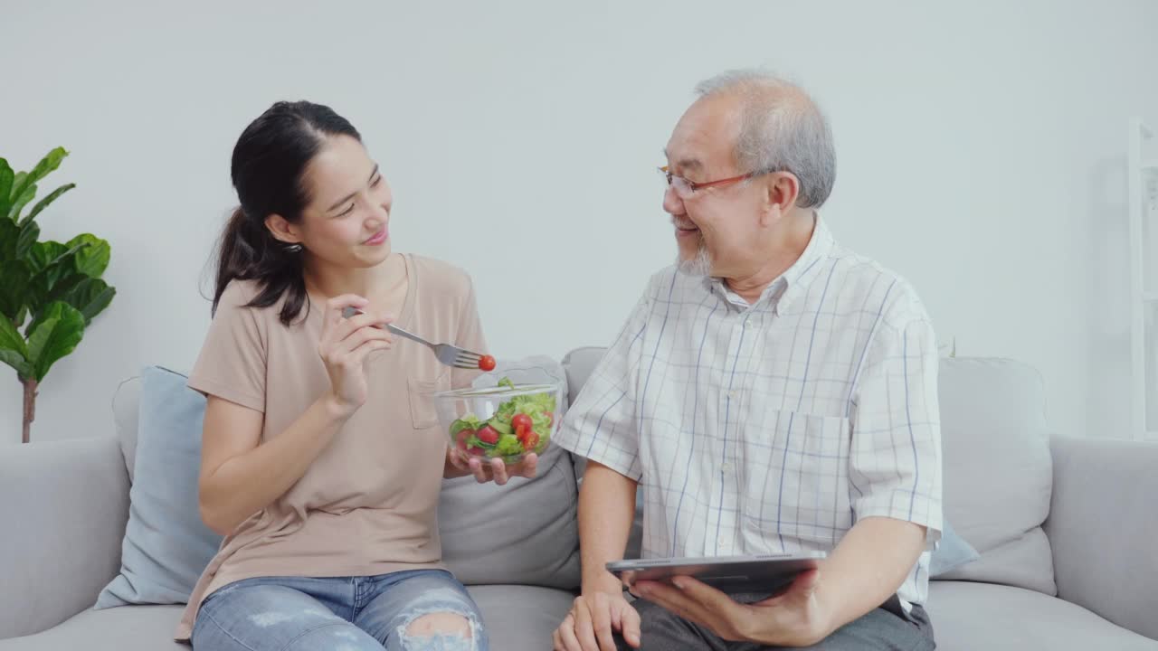 一只年轻的雌性正在给她的父亲吃蔬菜沙拉。父子俩愉快地坐在家里的沙发上度假。视频素材
