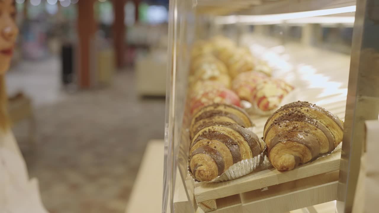 一位亚洲妇女正在看面包店货架上各式各样的羊角面包视频下载