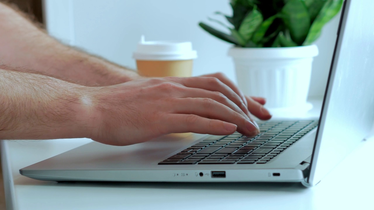 屋内光线明亮，男性的手正在笔记本电脑上打字，桌上放着一杯咖啡的一次性纸杯和一株绿色室内植物。办公室工作视频素材