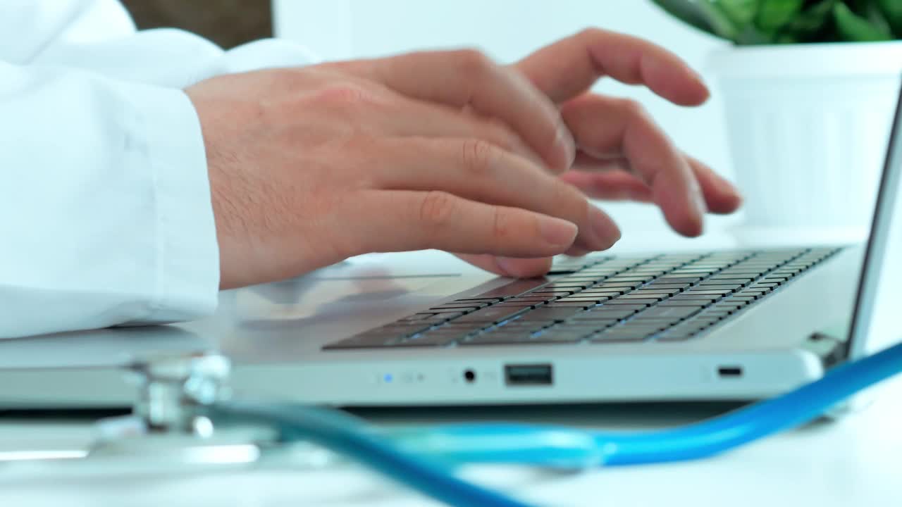 医学的概念，医生的工作，一个穿着白袍的男人的手在笔记本电脑上打印文本，在白色的桌子上一个风扇内窥镜和一株休眠的绿色植物视频素材
