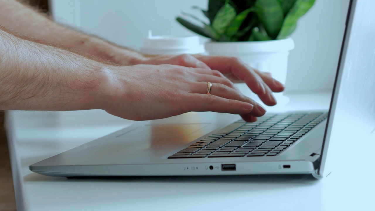 在一张白色的桌子上，男性用手在笔记本电脑键盘上打印文字，背景是一株绿色的室内植物。没有脸，只有特写视频素材