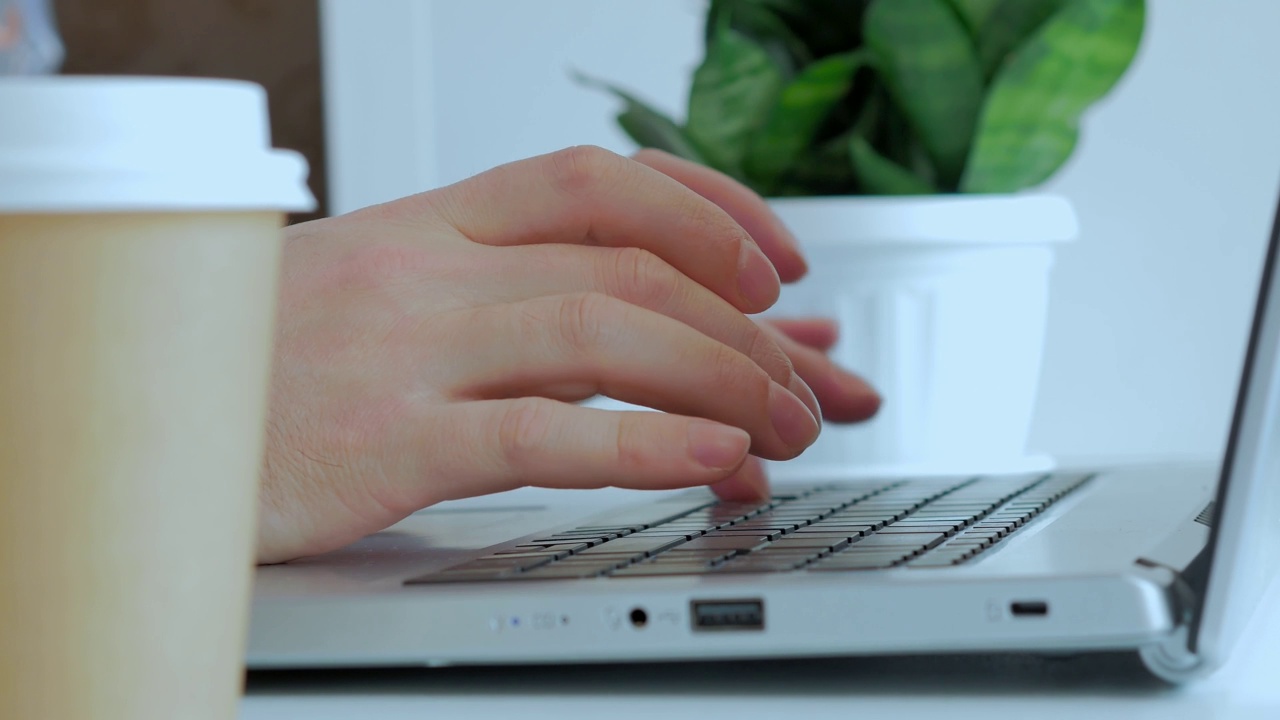 男性用手在白色桌子上的笔记本电脑键盘上打印文字，背景是一棵绿色的室内植物，还有一个装有咖啡的一次性纸杯。没有脸，只有特写视频素材