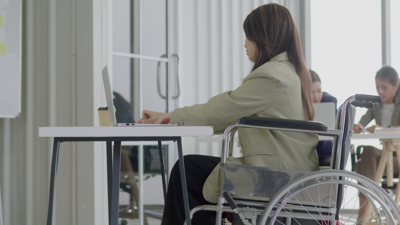 年轻的女商人坐在轮椅上，快乐地工作，平衡地工作和生活。坐在轮椅上的妇女——使用笔记本电脑在办公室工作。平等概念视频素材