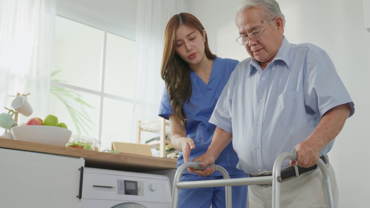 亚洲老年男性病人做物理治疗与照顾者。有吸引力的专业护理妇女帮助和支持年长的成熟男性实践行走缓慢与步行者在养老院照顾。视频素材