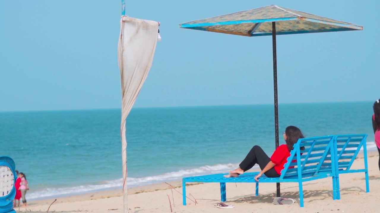 美丽的年轻印度女人在沙滩椅上放松。伞椅和海滩的侧视图。暑假的背景。在印度古吉拉特邦的Shivrajpur海滩。度假和旅行的概念。视频素材